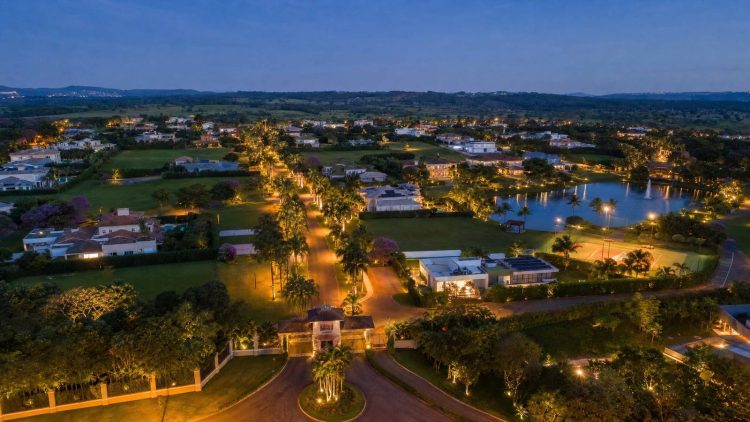 Vista aérea de condomínio de lotes de alto padrão ao entardecer, com lotes amplos, vias internas arborizadas, lago central e portaria