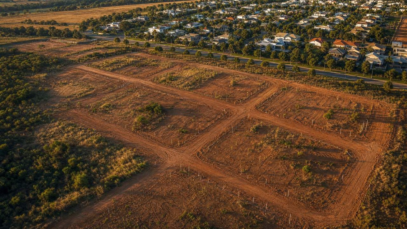 Vista aérea de loteamento irregular ao entardecer, com lotes apenas demarcados em terra, sem pavimento nem infraestrutura, ao lado de um bairro regularizado